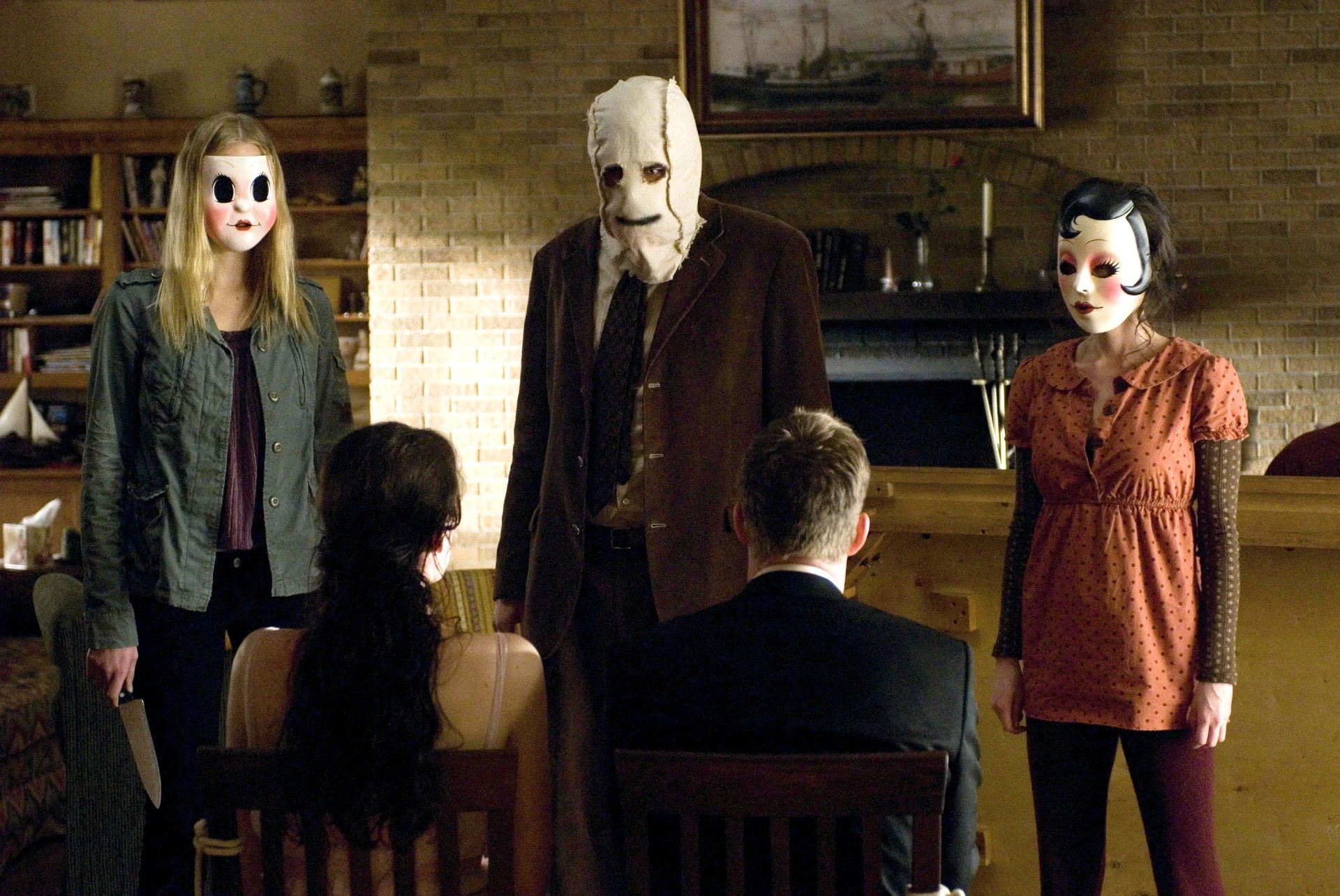 Three masked figures stand in front of a couple in The Strangers.