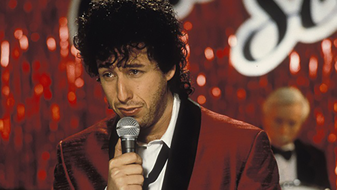 Adam Sandler com cabelos cacheados segurando um microfone e cantando em cena de The Wedding Singer.