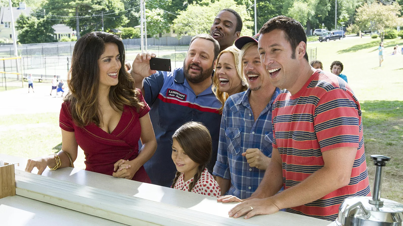 Een groep mannen, vrouwen en kinderen, waaronder Adam Sandler en Salma Hayek, staan ​​aan de balie, lachend en glimlachend in Grown Ups.