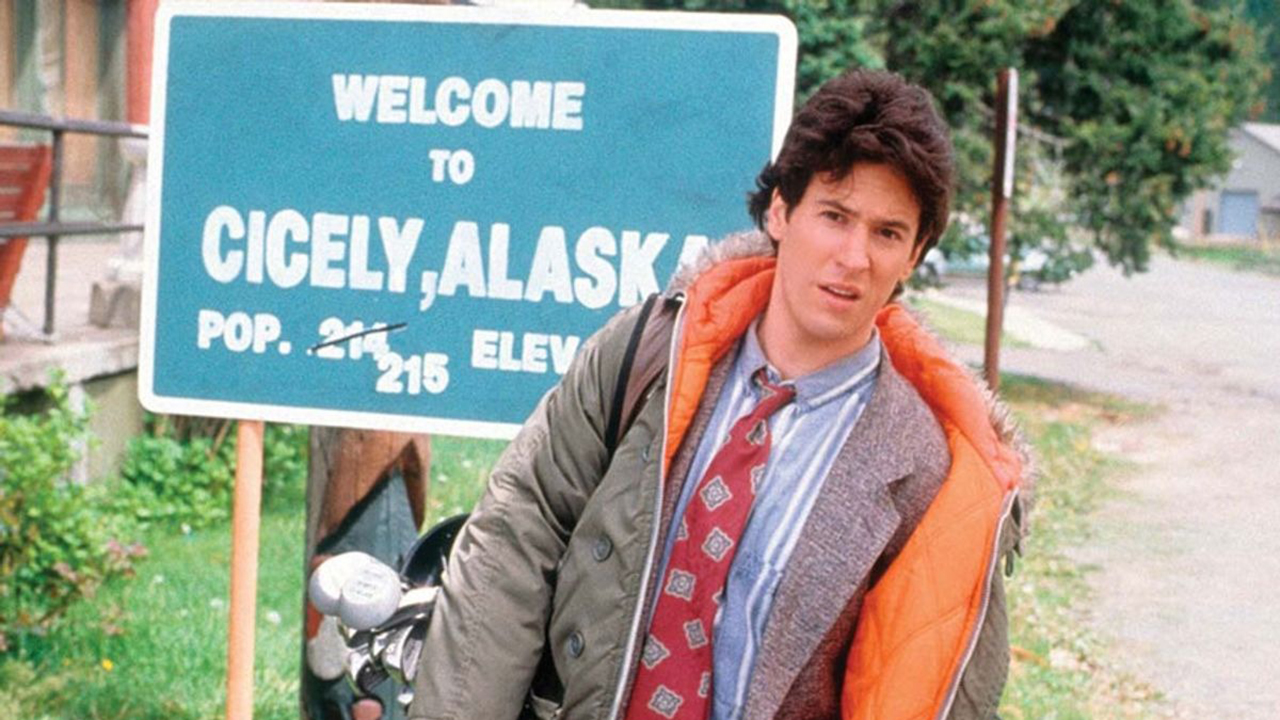 Joel parado em frente a uma placa que diz bem-vindo a Cicely, Alasca, em uma cena de Northern Exposure.