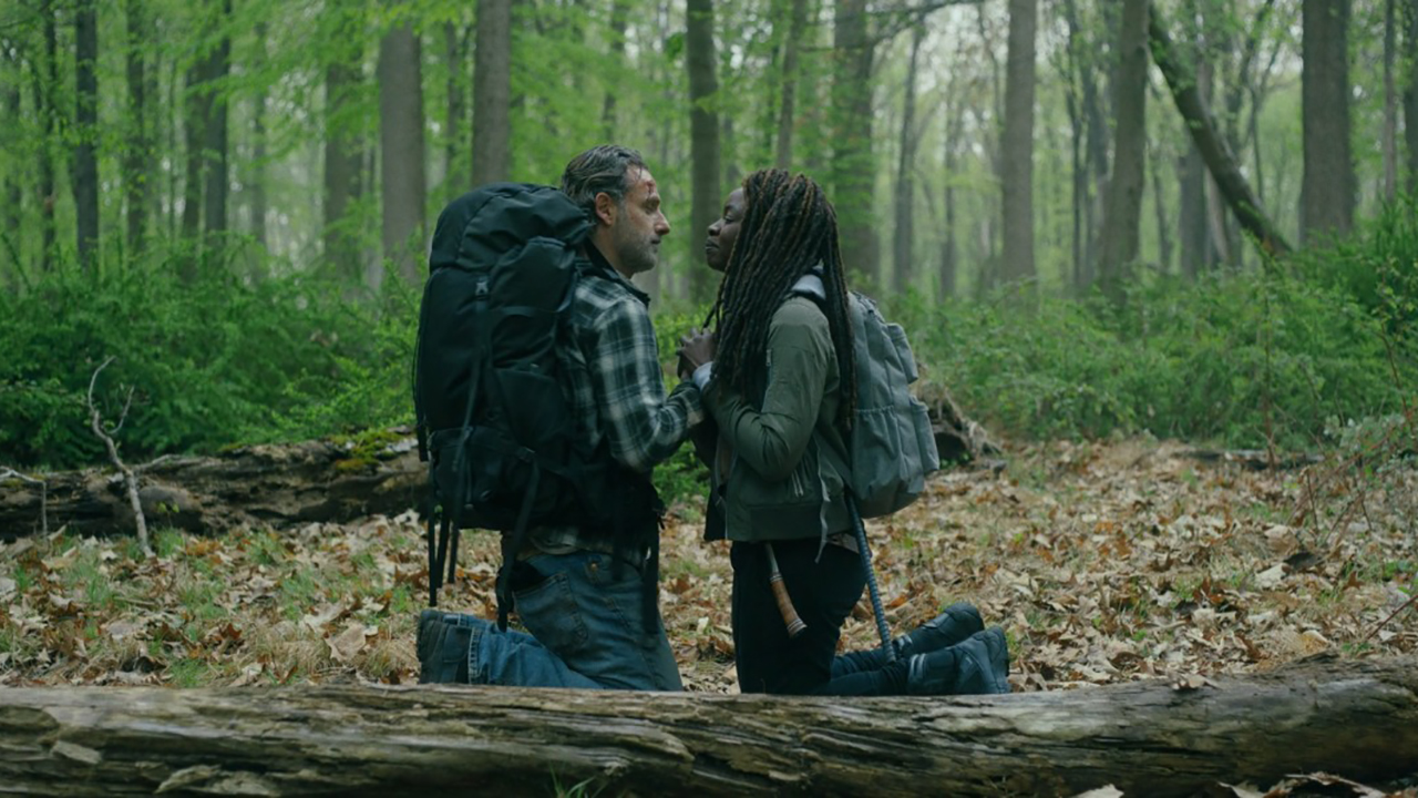 Rick e Michonne à paisana com mochilas, ajoelhados e frente a frente em uma floresta em The Walking Dead: The Ones Who Live.