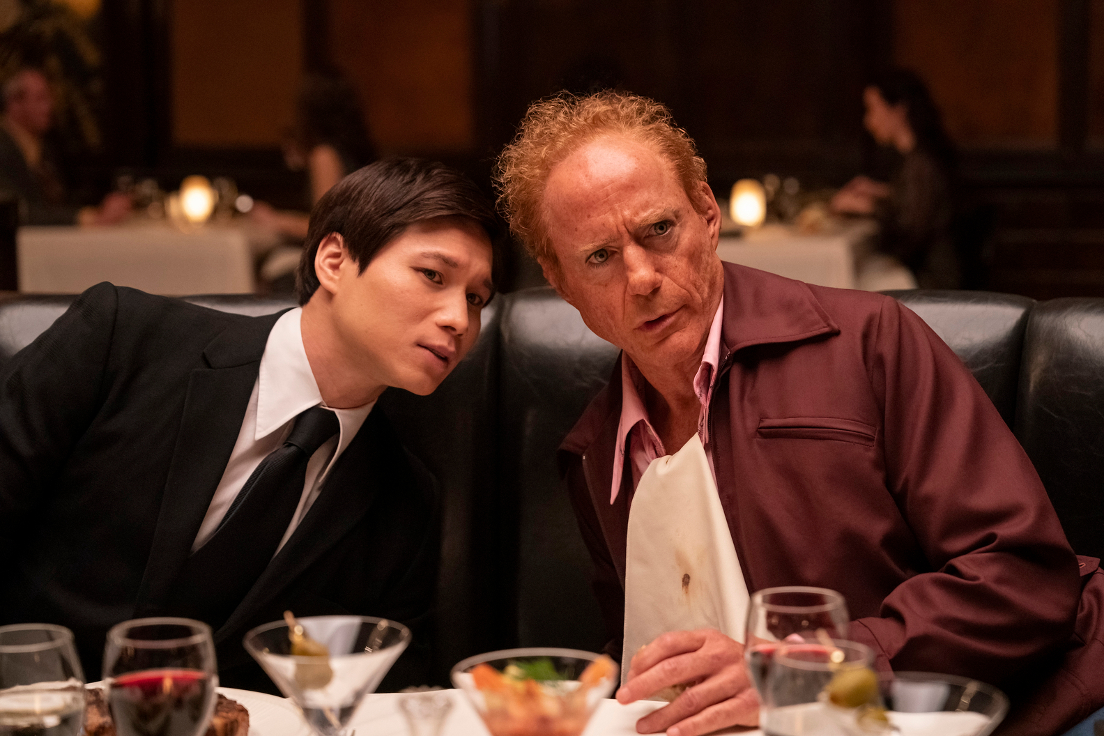 Hoa Xuande and Robert Downey Jr. sit at a dinner table together in The Sympathizer.