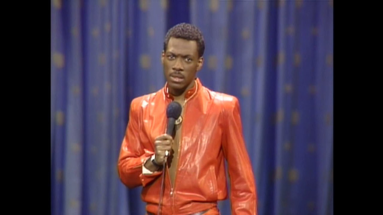 Eddie Murphy fazendo uma careta no icônico terno de couro vermelho enquanto está no palco com um microfone em Eddie Murphy Delirious.