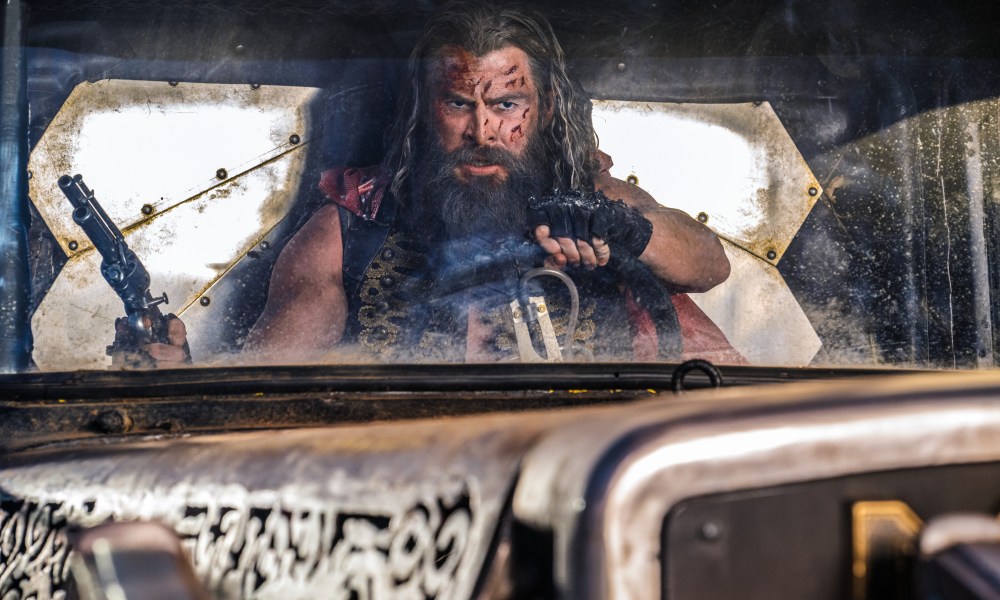 A bloodied Chris Hemsworth holds a gun and a steering wheel in Furiosa: A Mad Max Saga.