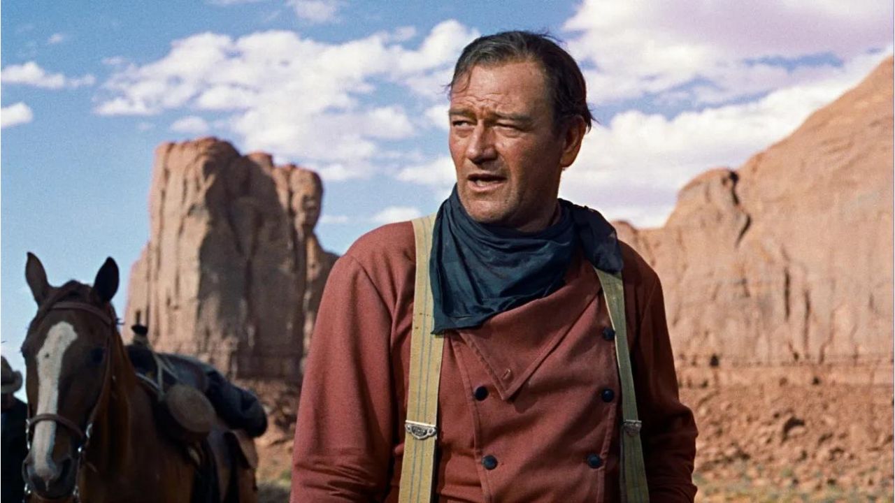 John Wayne como Ethan Edwards en el desierto mirando atentamente algo fuera de cámara en The Searchers.