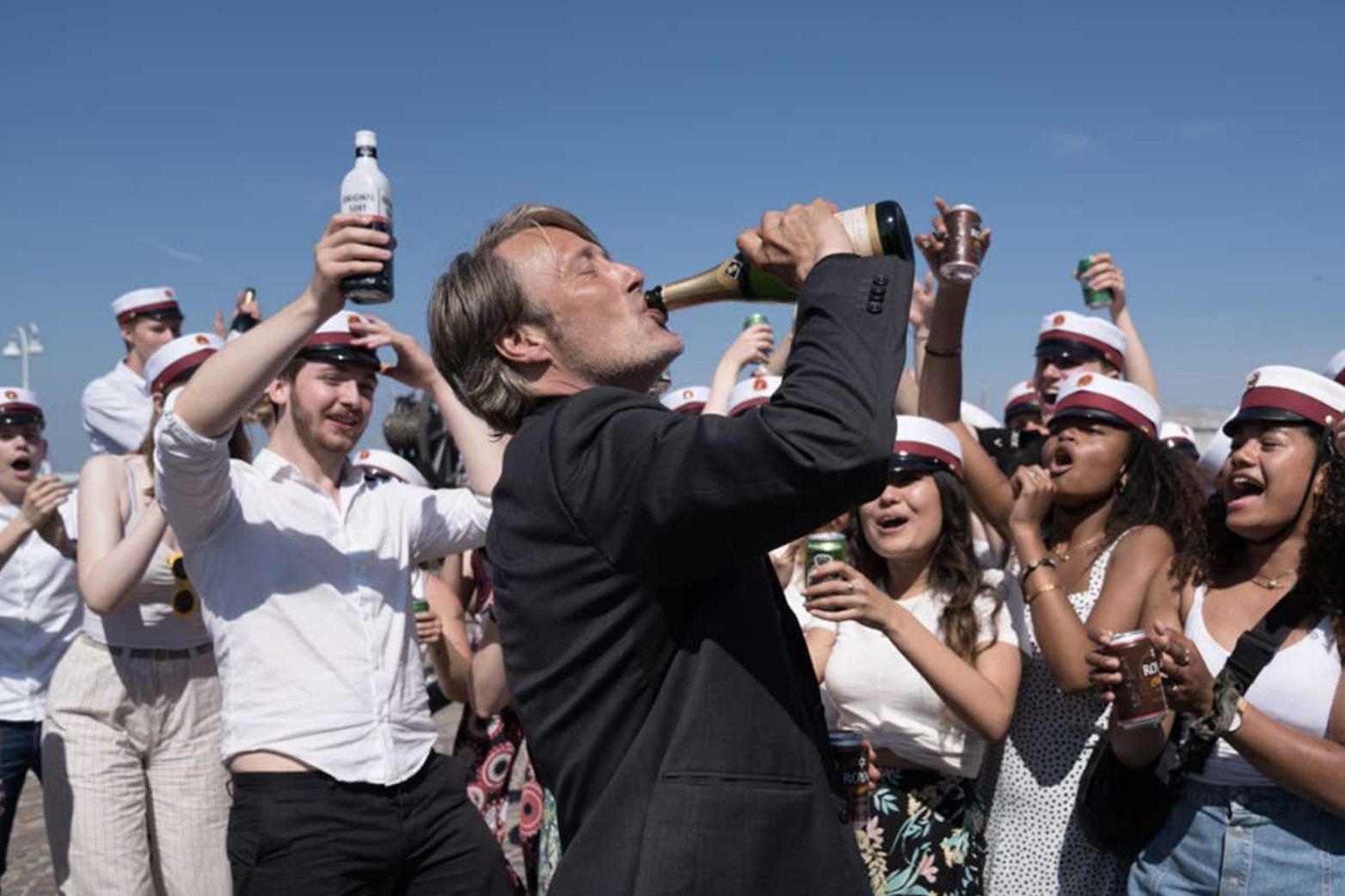 A man drinks a bottle of champagne with people looking on behind him.