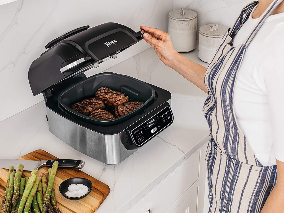 A woman looks at the <a href='https://ai-blog.writing-club.top/tag/business' target='_blank'>food</a> cooking in the Ninja Foodi 5-in-1 indoor electric grill.”><figcaption id=