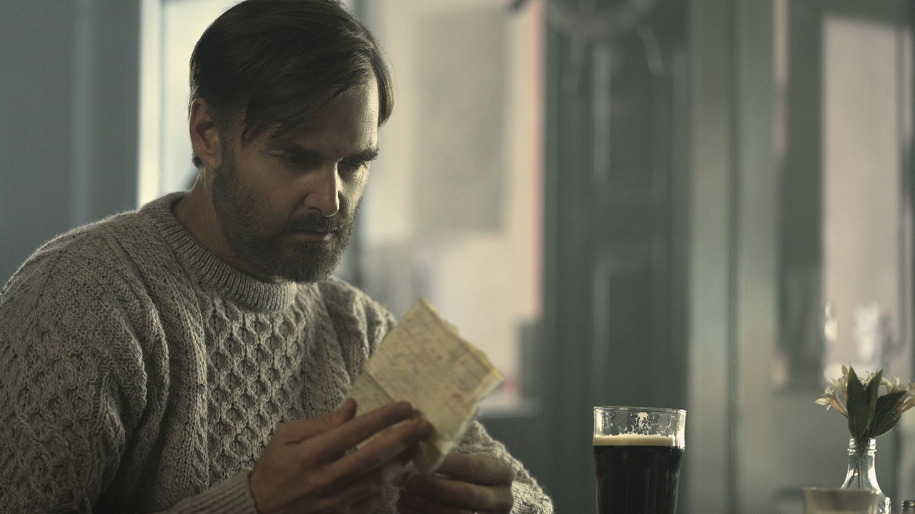 Will Forte sitting at a desk looking at a paper, a beer beside him in a scene from Bodkin.