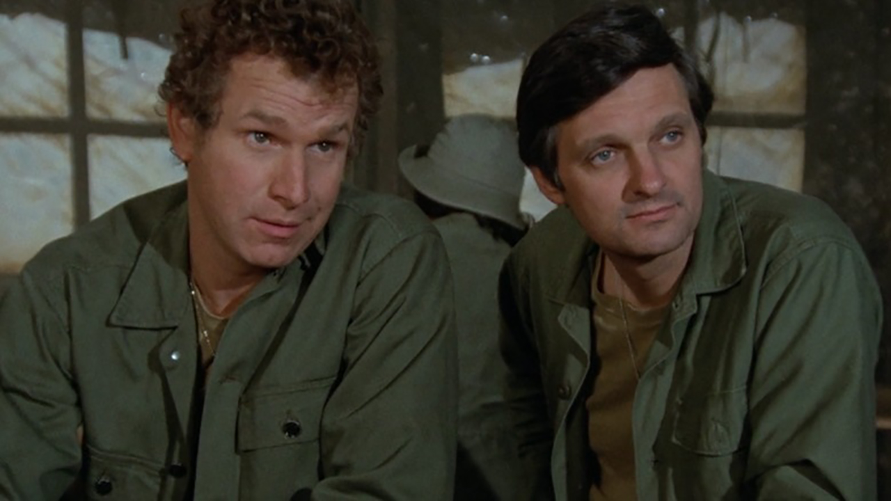 《M*A*SH》的场景中，两个男人坐在一起，都穿着绿色军用外科制服。