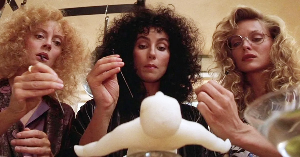 Three women hover over a voodoo doll in The Witches of Eastwick.