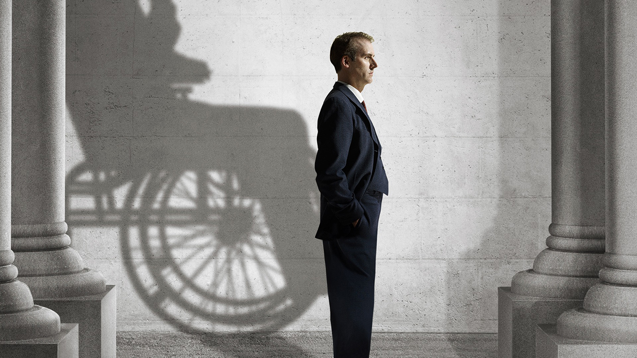 Un homme debout en costume tourné vers la droite, une silhouette d'une personne en fauteuil roulant derrière lui de la mini-série The World Wars.