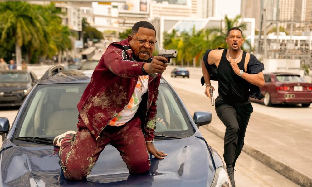 Martin Lawrence sits on a car hood while Will Smith runs next to him in Bad Boys: Ride or Die.