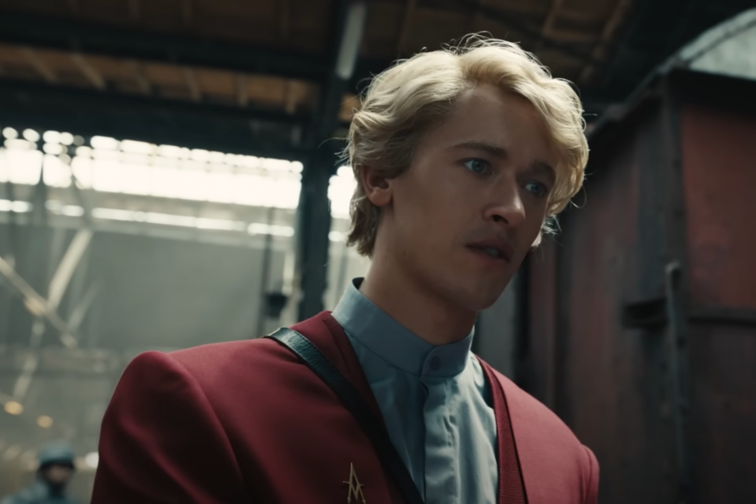 A blond man in a red suit stands and stares.