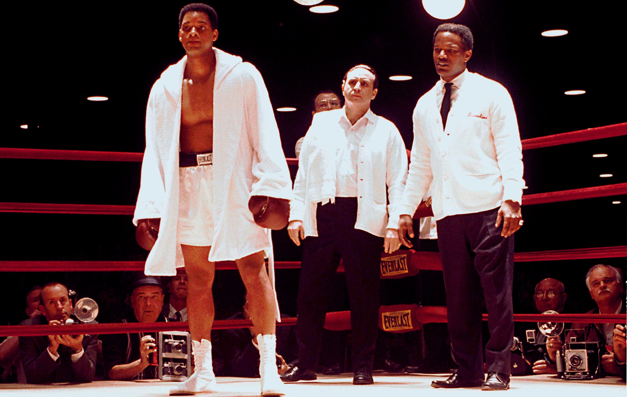 अली में एक बॉक्सिंग रिंग में तीन आदमी खड़े हैं।