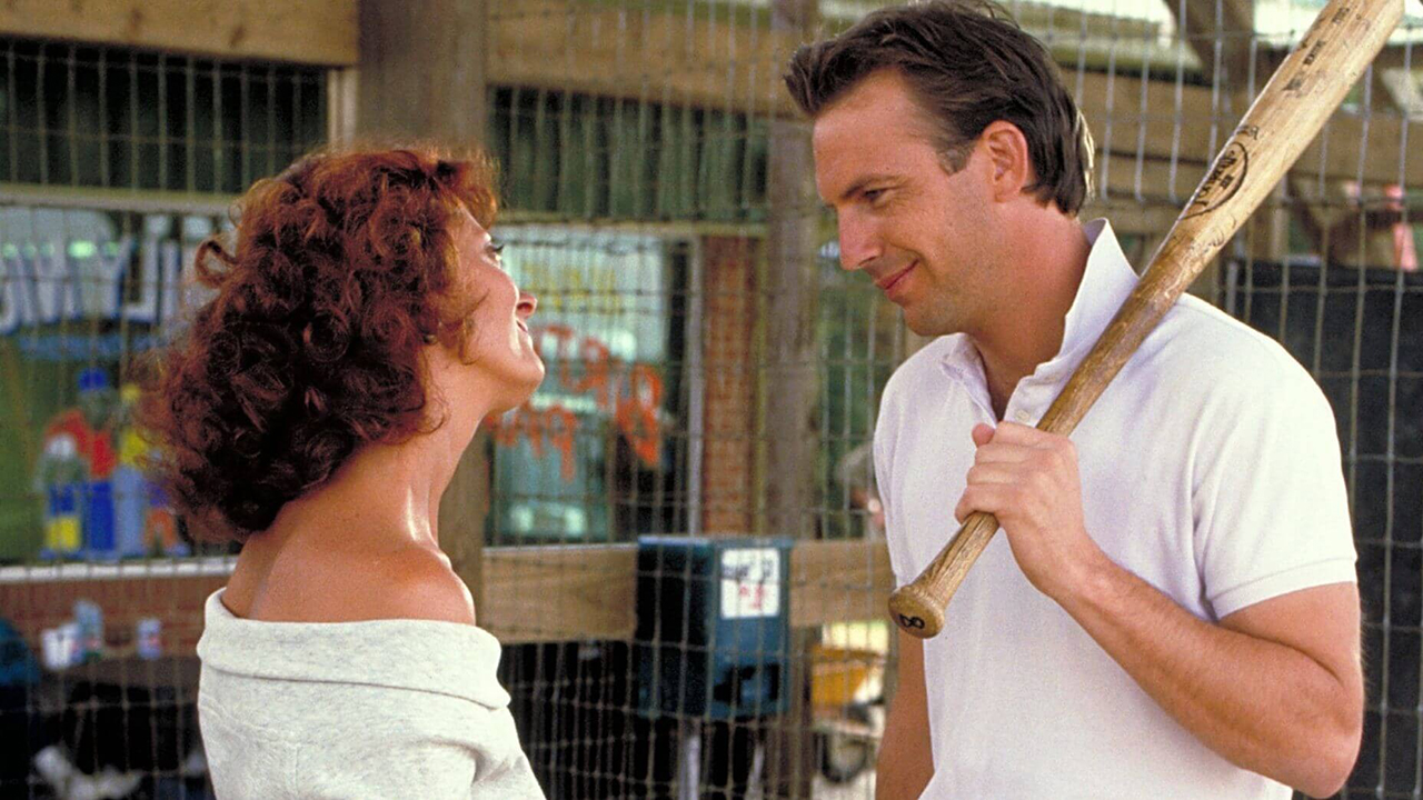 Susan Sarandon schaut liebevoll Kevin Costner an, der in einer Szene aus Bull Durham einen Baseballschläger hält.