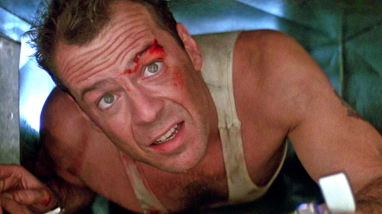 Bruce Willis crawling under something in a tank top with a lighter in a scene from Die Hard.