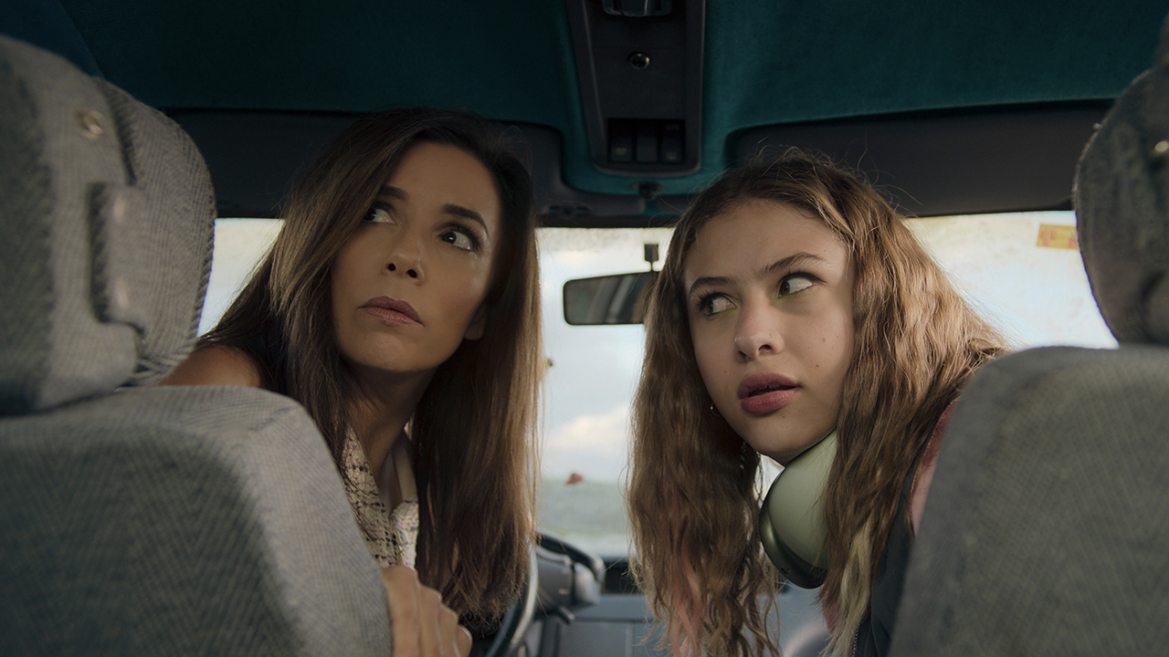 Eva Longoria is seen on a plane, with a young teenager next to her. They both look back in a scene from the film Womanland on Apple TV+.