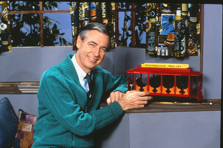 弗雷德·罗杰斯 (Fred Rogers) 在罗杰斯先生街区的手推车长凳旁摆姿势。