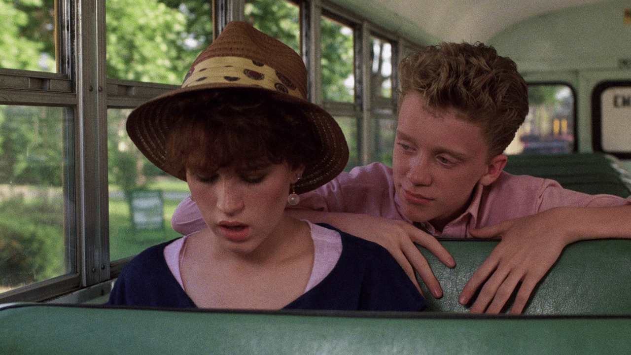 Molly Ringwald wearing a hat sitting on a school bus, a young boy looking over her shoulder from the seat behind her in Sixteen Candles.