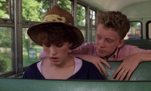 Molly Ringwald wearing a hat sitting on a school bus, a young boy looking over her shoulder from the seat behind her in Sixteen Candles.