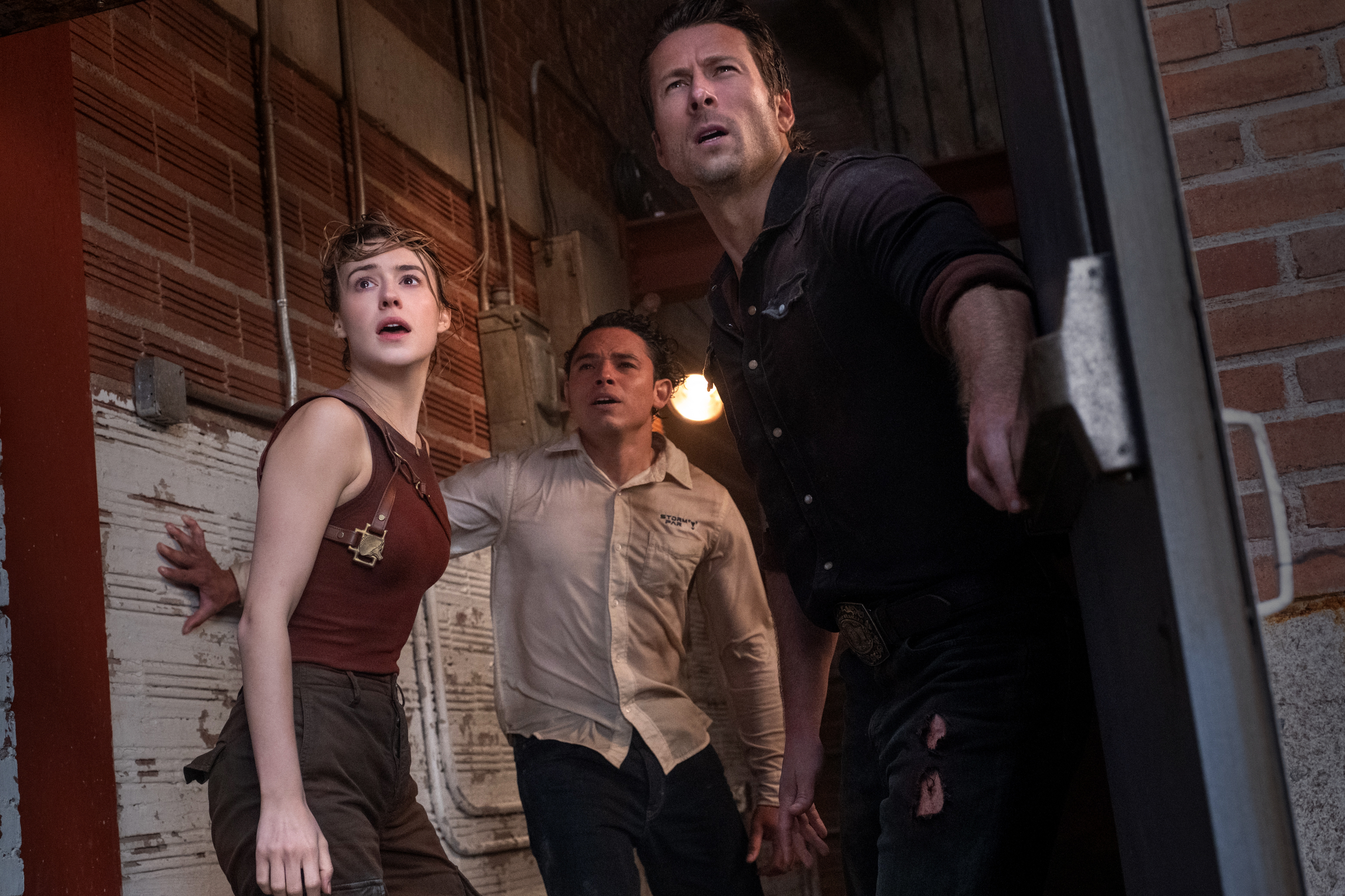 Daisy Edgar-Jones, Anthony Ramos, and Glen Powell look out an open doorway in fear in Twisters.