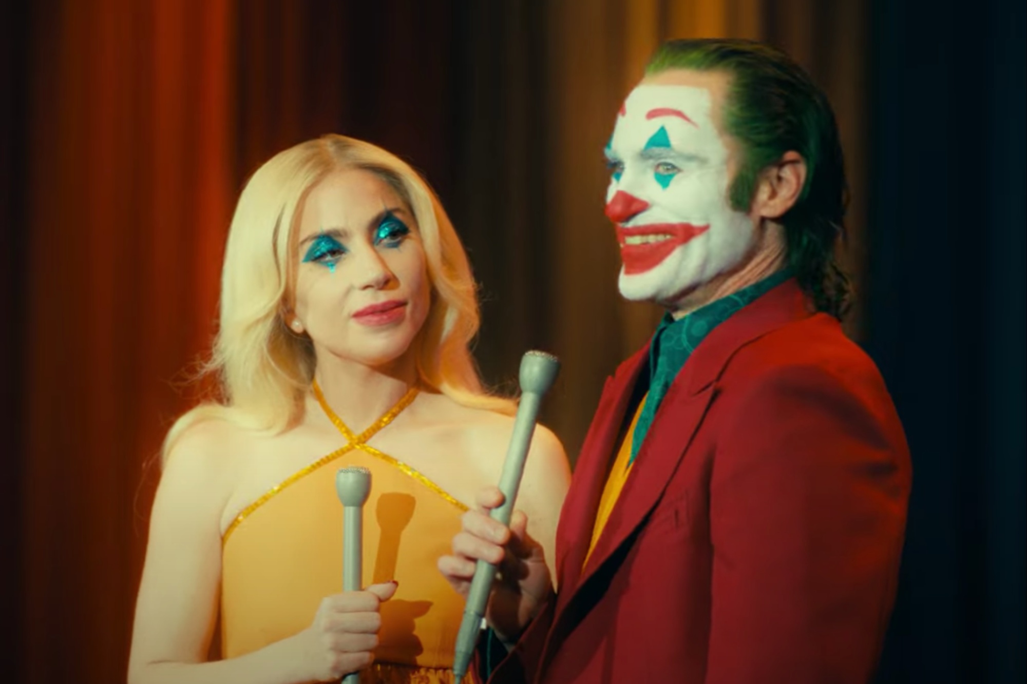 Lady Gaga y Joaquin Phoenix sostienen micrófonos y sonríen en Joker 2.