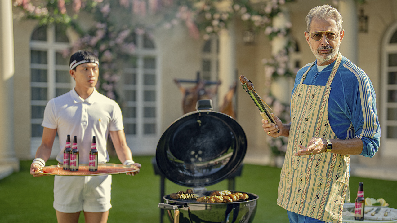 Jeff Goldblum barbecuing with an apron, another man holding a tray beside him in a scene from Kaos on Netflix.