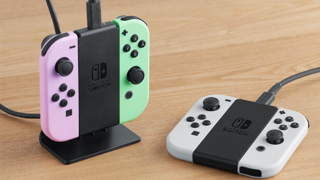 Joy-Cons violets et verts sur le support de chargement noir. Il y a un autre support de chargement à côté, mais il repose à plat sur la table avec deux Joy-Cons blancs.