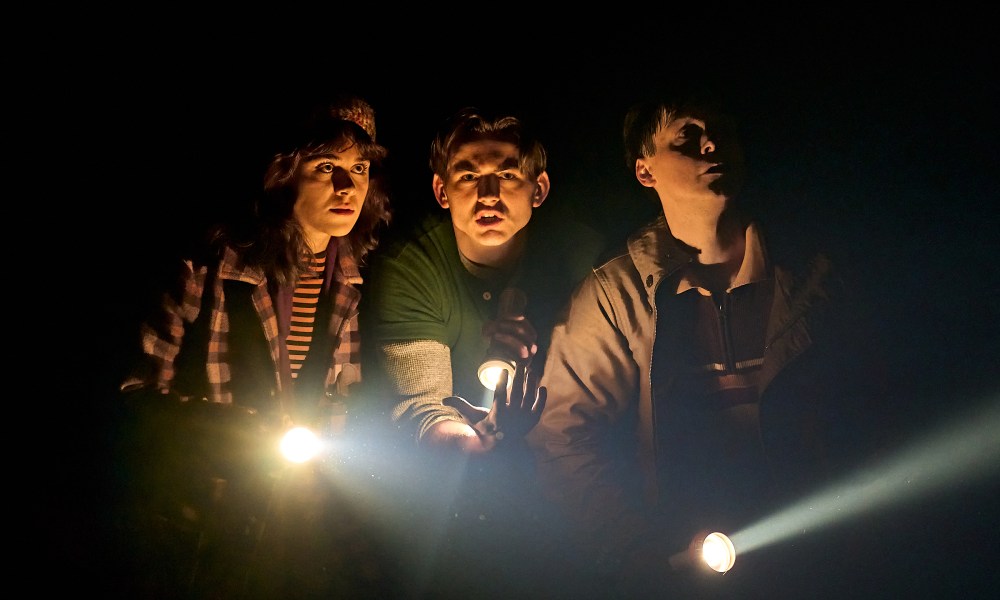 Two boys and one girl stand next to each other in the dark while holding flashlights.