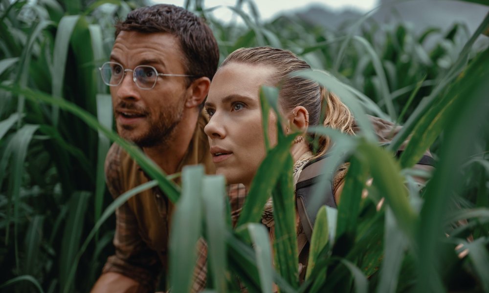 A man and woman hide in a field and stare curiously.