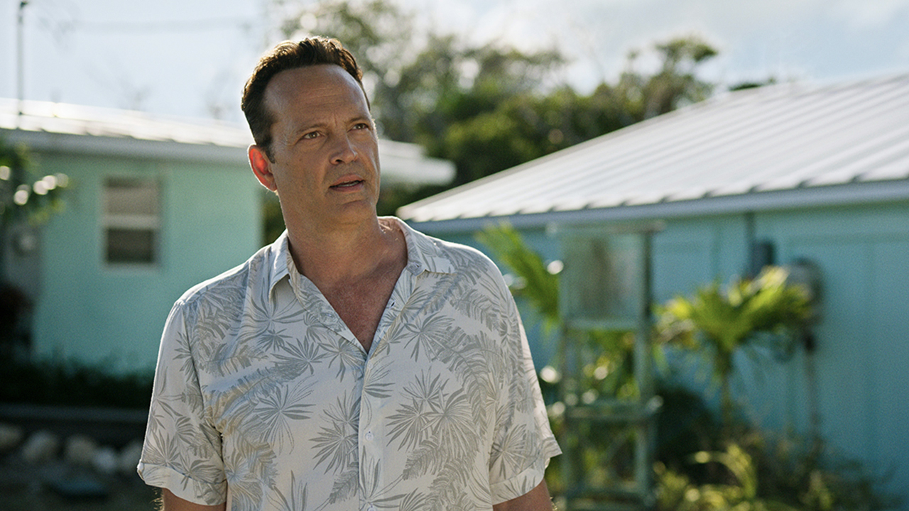 Vince Vaughn in a floral button top standing by a beach house looking annoyed in a scene from Bad Monkey.