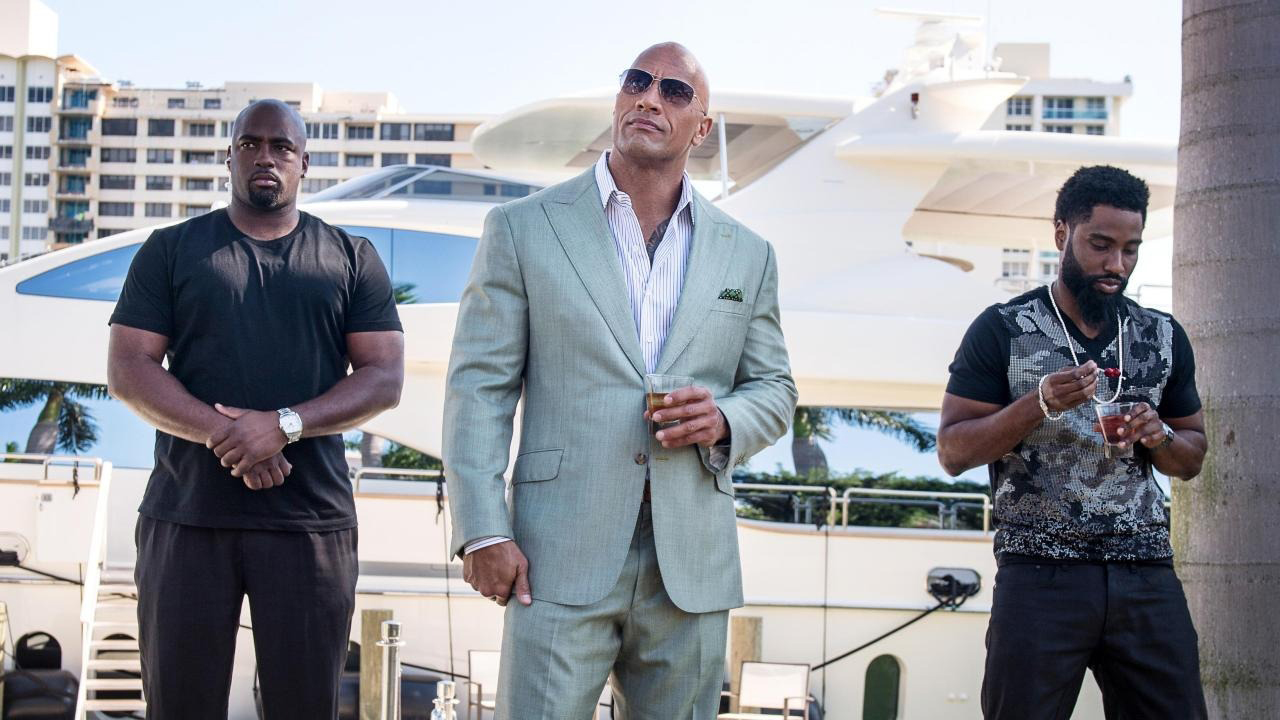 Dwayne "The Rock" Johnson in a blue suit wearing sunglasses with two men on either side in a scene from Ballers.