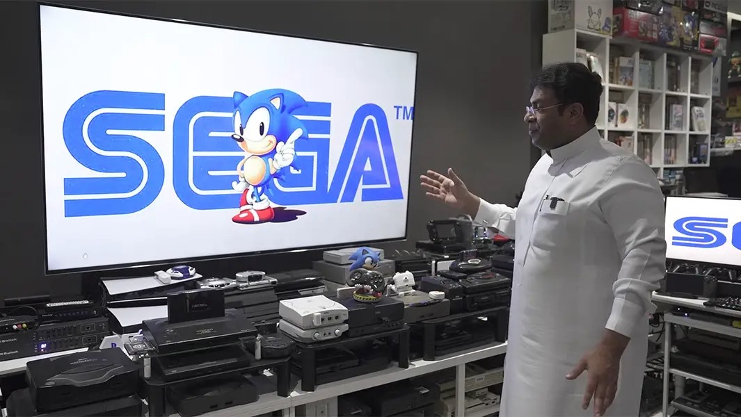 Ibrahim Al-Nasser standing in front of his console setup. There are a ton of consoles lined up in front of a TV. Sonic and the Sega logo are on the TV