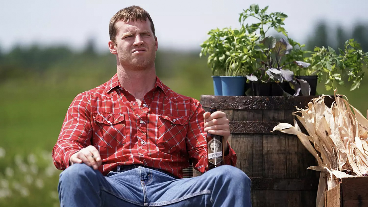 Um homem sentado do lado de fora com uma camisa xadrez vermelha segurando uma cerveja e apertando os olhos sob o sol em Letterkenny.