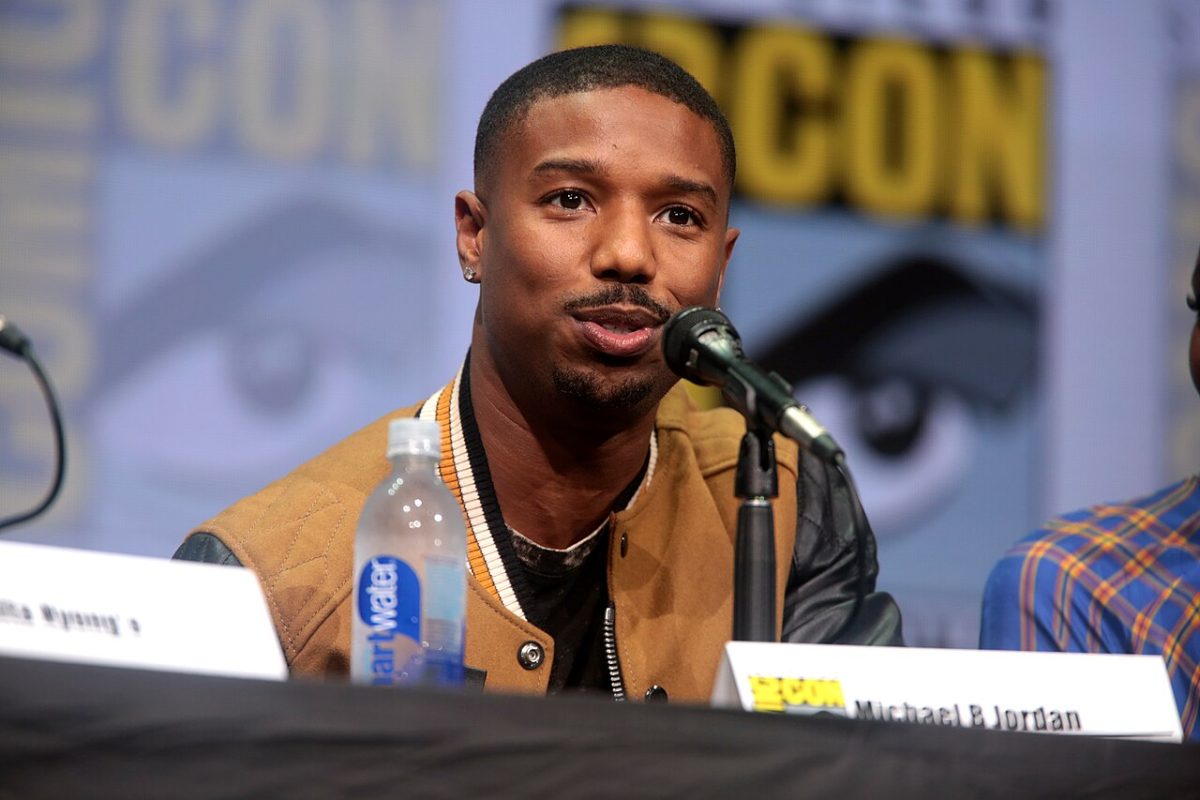 迈克尔·B·乔丹 (Michael B. Jordan) 坐在讲台上讲话。