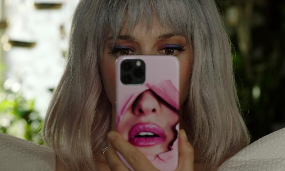 A young woman with pink hair peers at her pink phone that shows lips pushing through paper in Under the Influencer.