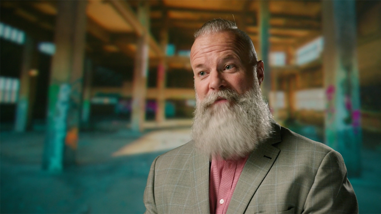 A man with a long beard sits on a chair in an empty room and is interviewed in “Caught at Last.”