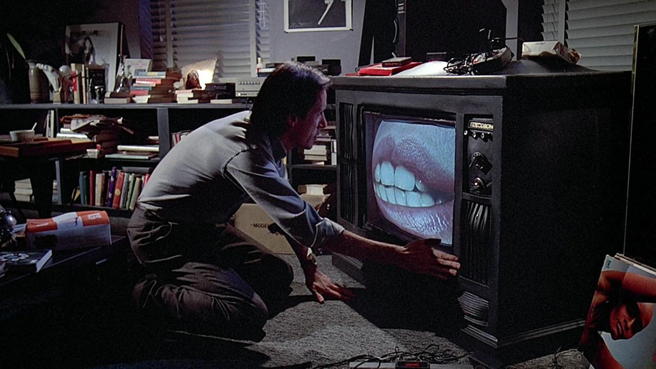 Um homem agachado em frente a uma TV de tubo com um par de lábios ampliados na tela em cena do Videodrome.