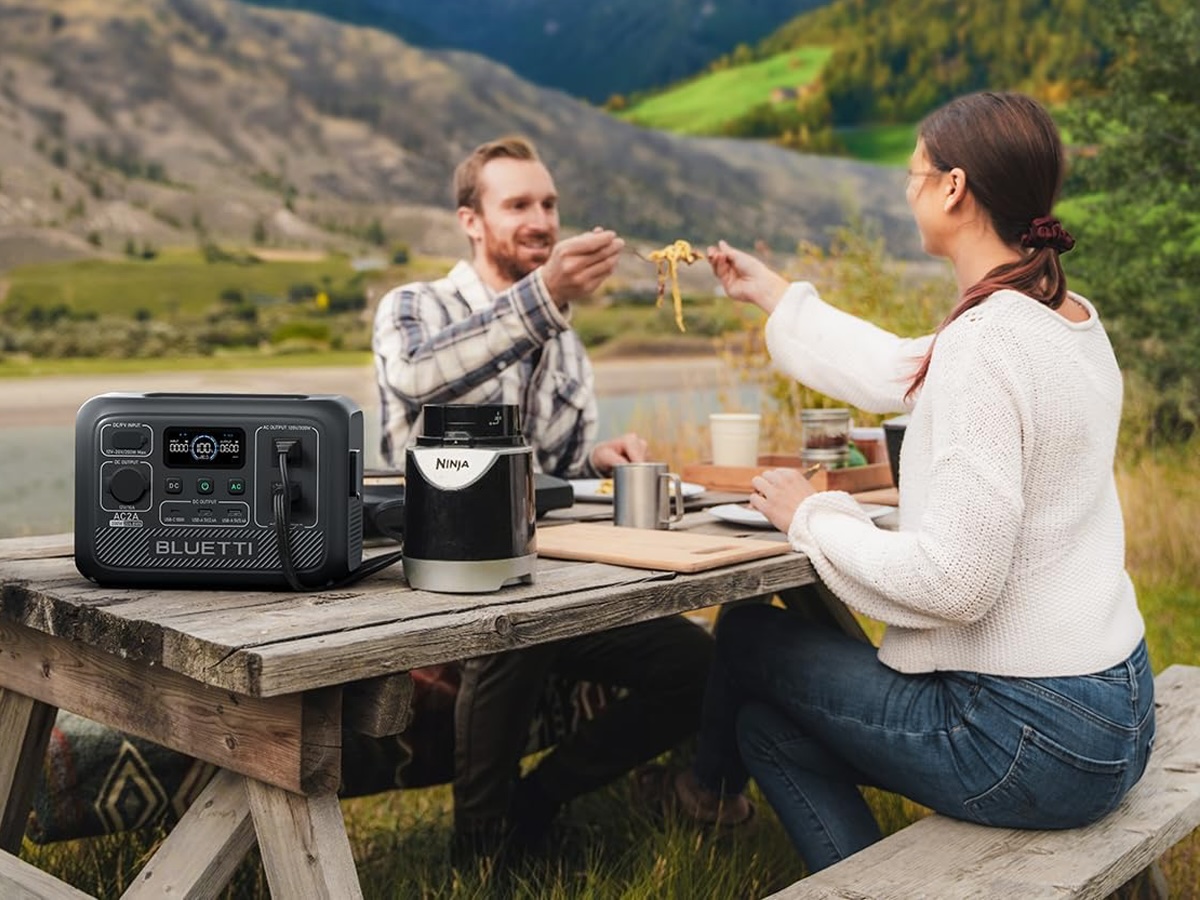 Ein Paar isst beim Camping eine Mahlzeit mit der tragbaren Bluetti AC2A-Stromstation auf dem Tisch neben ihnen.