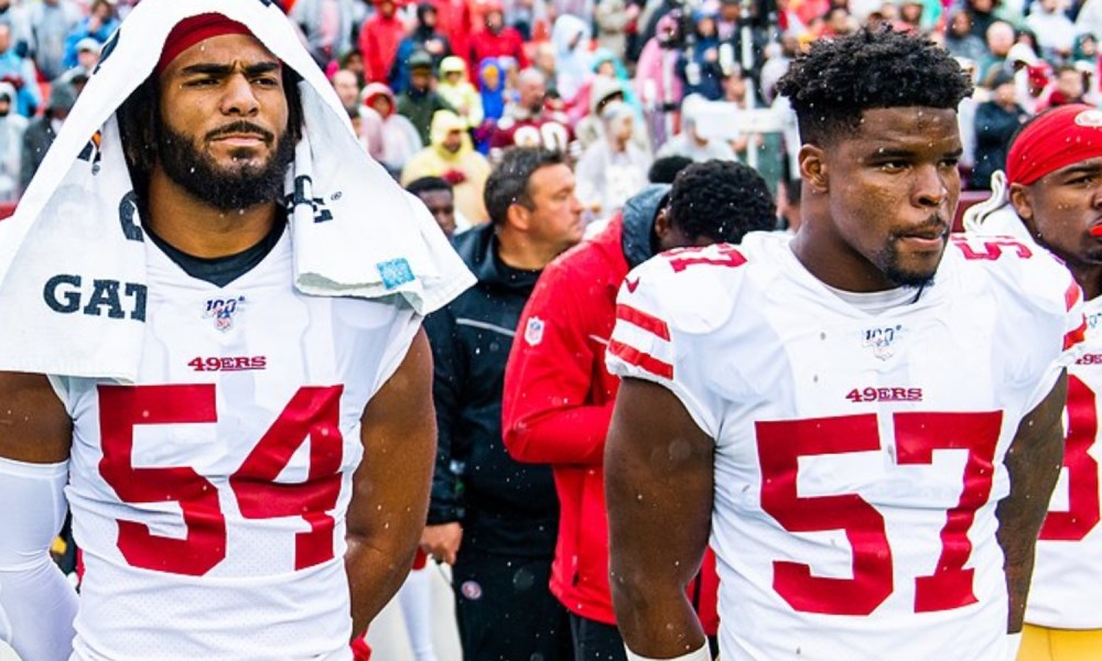 Two linebackers stand next to each other on the sidelines.