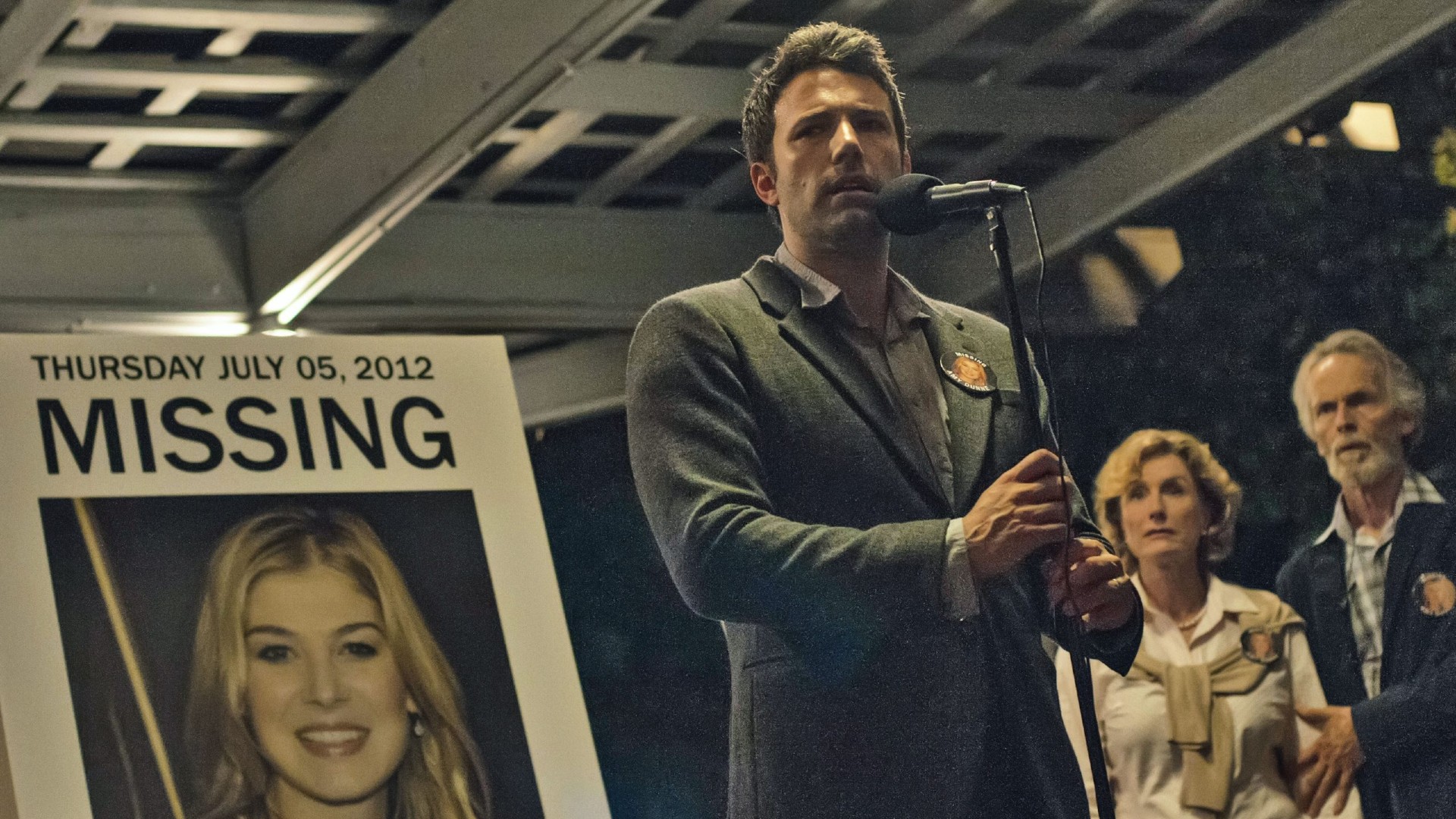 Ben Affleck está em frente a uma foto ampliada de Rosamund Pike, segurando um microfone, em uma foto do filme Gone Girl.