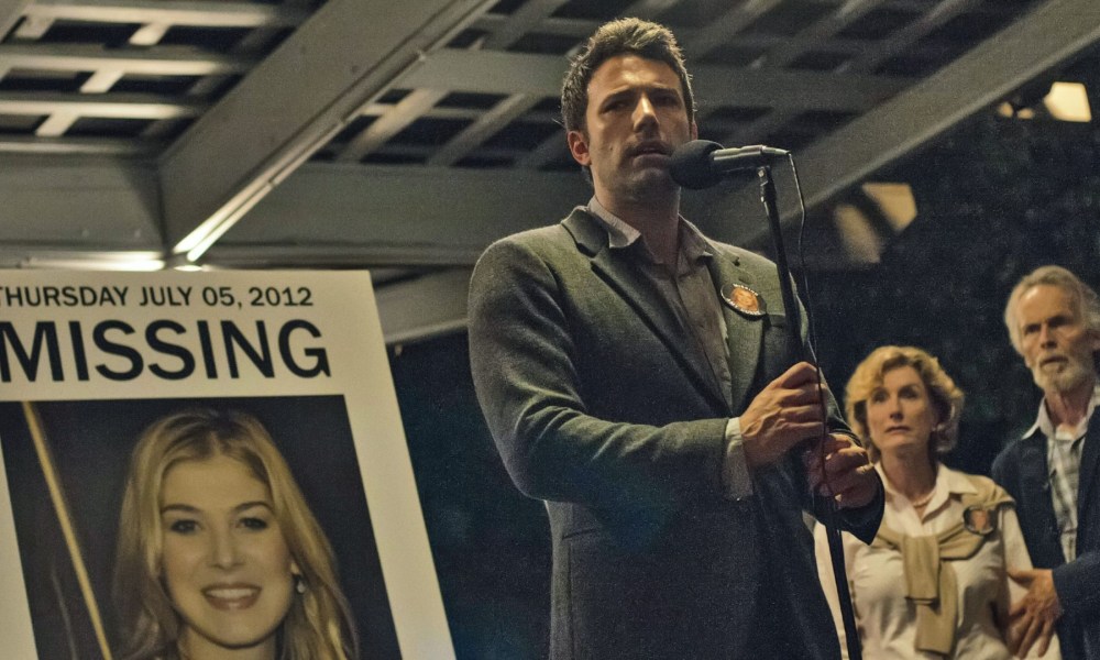 Ben Affleck stands in front of a blown-up photo of Rosamund Pike, holding a microphone, in a still from the movie Gone Girl.