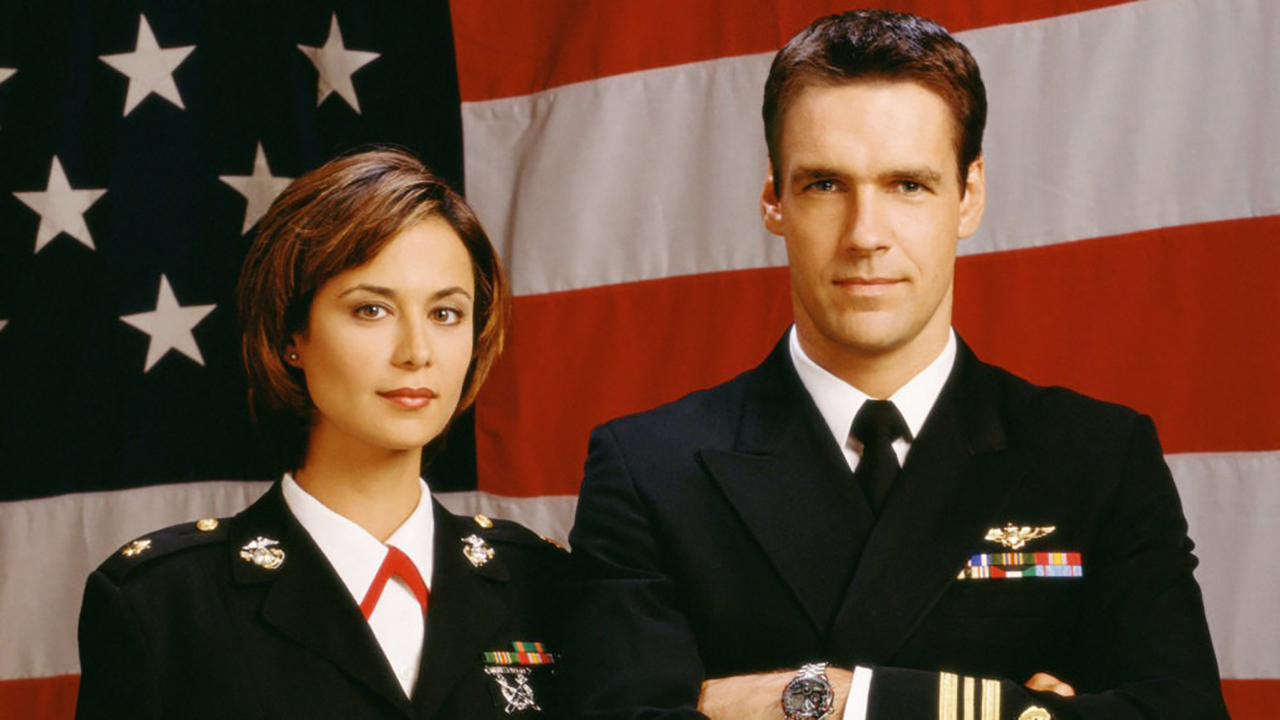 A woman in man in military uniforms standing in front of an American flag in JAG.