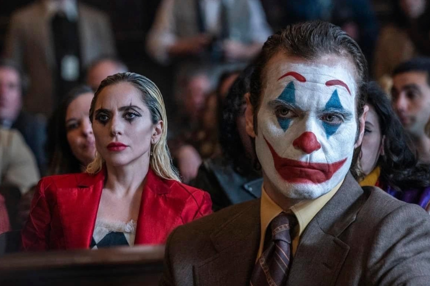 Joker bir masada oturuyor, Harley Quin ise arkasında oturuyor.