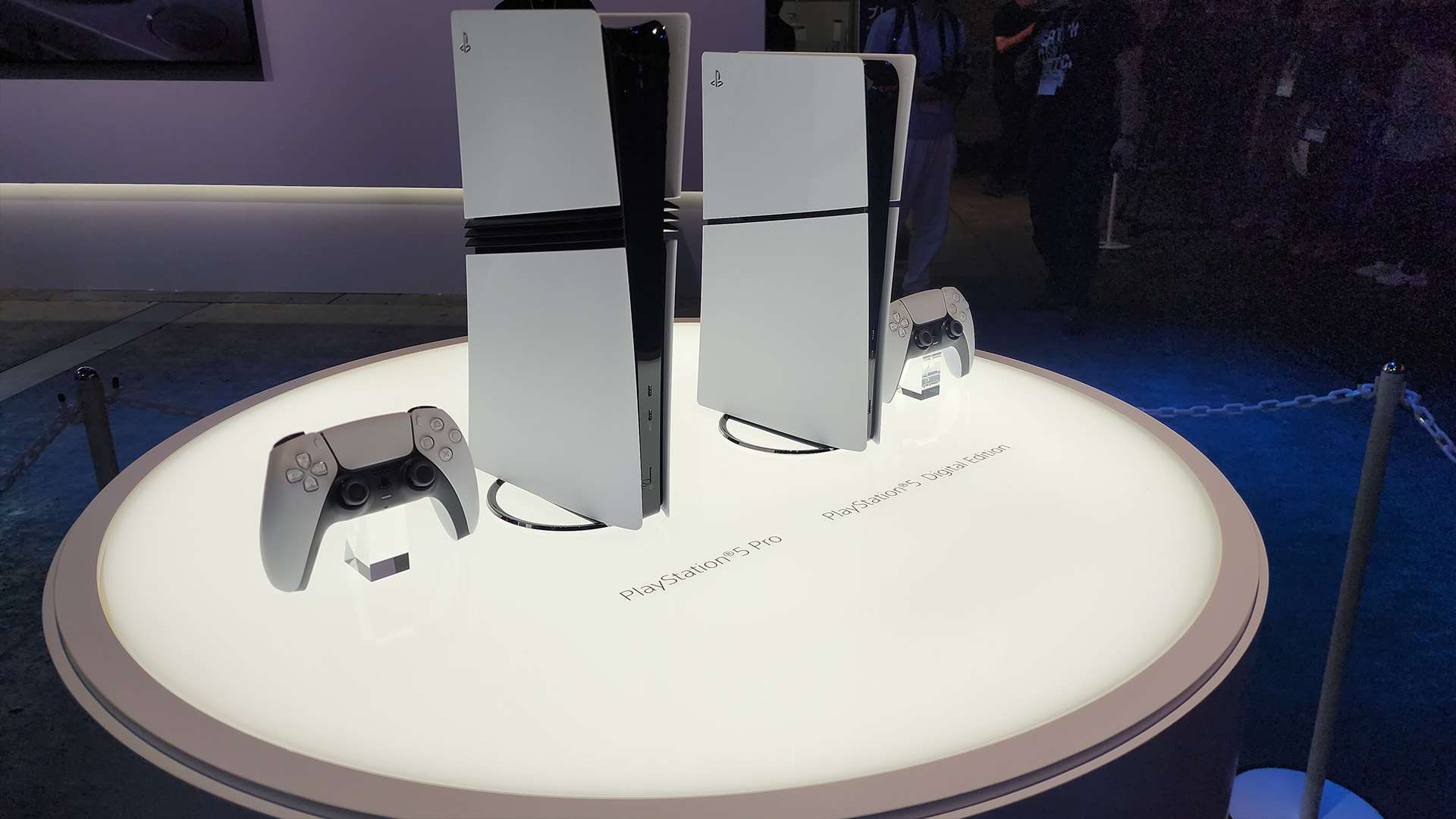 A lit table holds a PS5 Pro and an all-digital PS5, the former being noticeably taller.