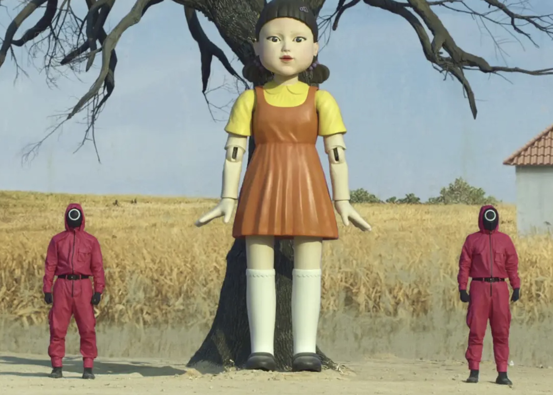 A giant doll stands in the middle of two men with red jump suits.