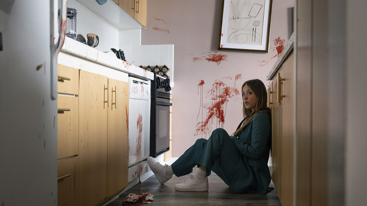 Ava is sitting on the floor of a kitchen, with blood splattered on the path behind her, in Based on a True Story.