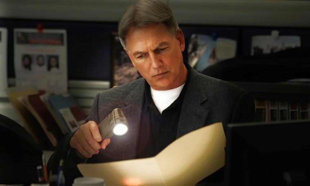 Mark Harmon holding a flashlight up to documents and looking intently at them in NCIS.