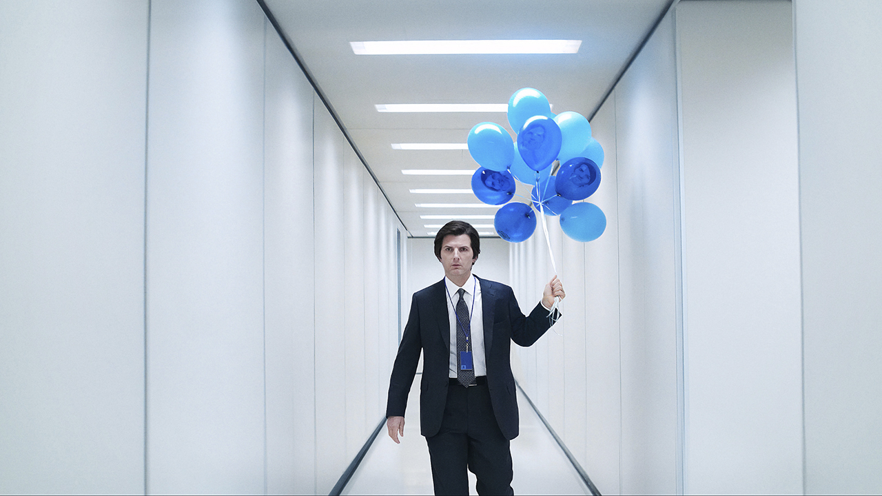 Mark walking down the bright white hallway at work looking angry, carrying a bunch of bright blue balloons in Severance.