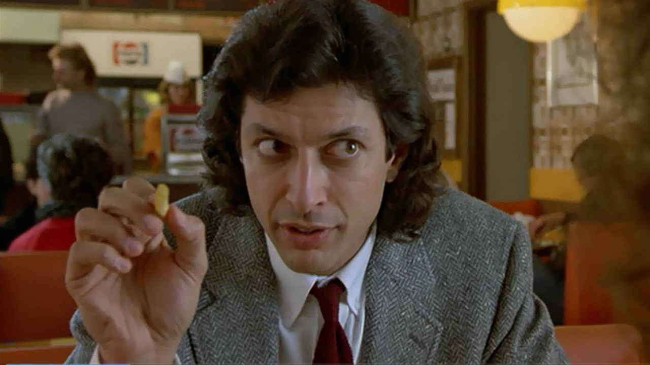 Jeff Goldblum en costume tenant un morceau de nourriture dans un restaurant de The Fly.